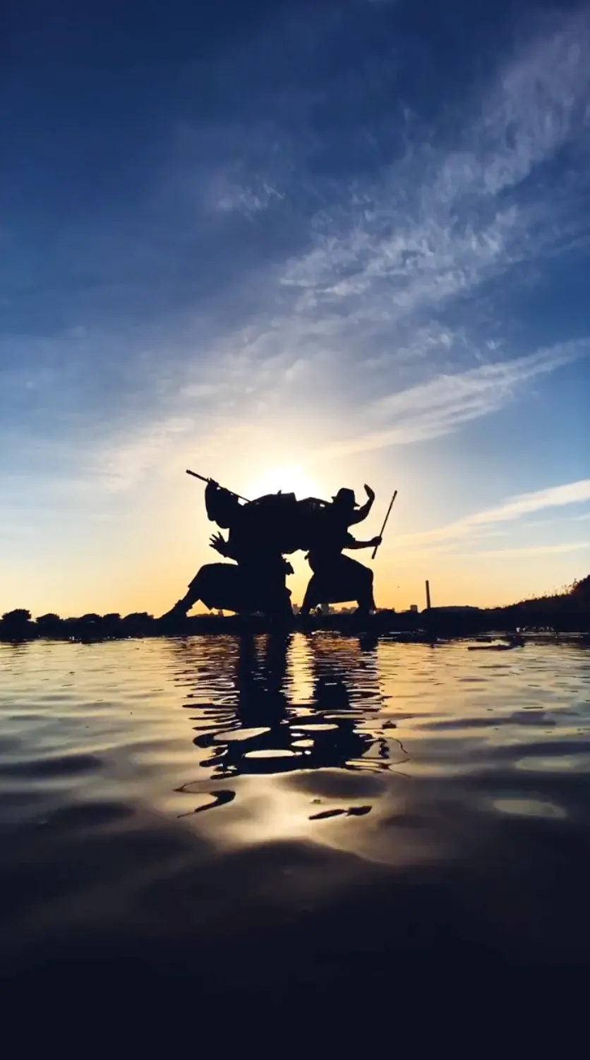 夕日を背景に、水辺で対峙する二人の武士のシルエット。刀を構えた姿が水面に映り込んでいる