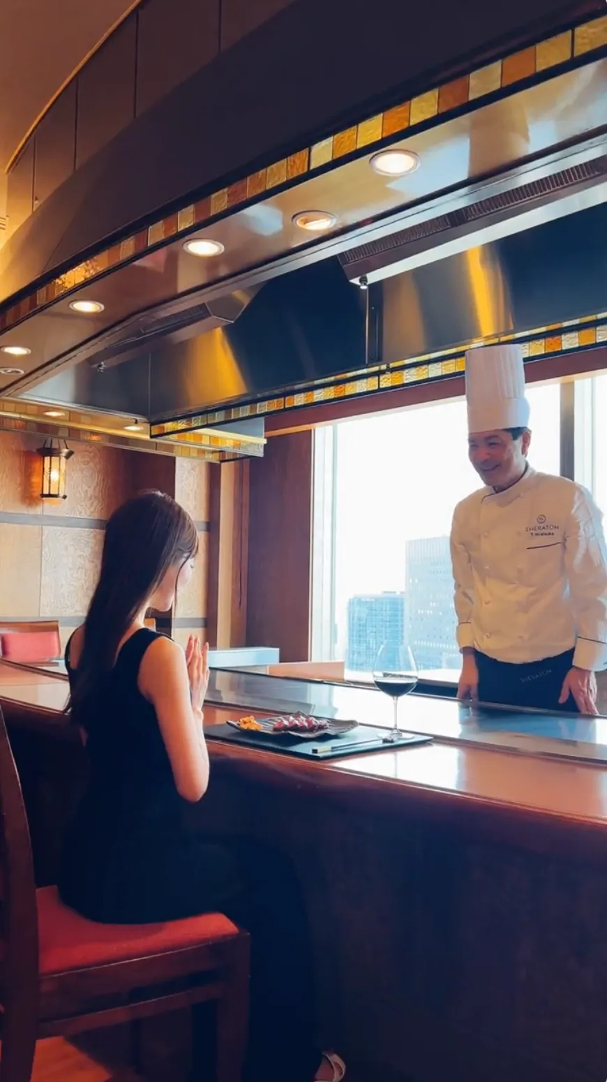横浜ベイシェラトンのレストランで、シェフが笑顔で料理を提供し、女性ゲストが料理を楽しみに待つ様子を映した写真。窓の外には横浜の街並みが広がり、ホテルならではの上質な空間と食の体験が表現されている。